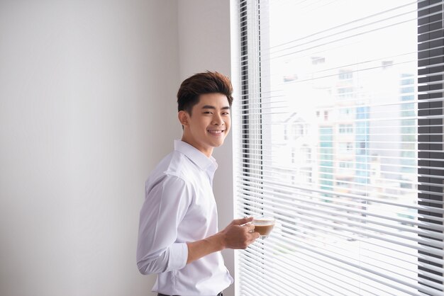 Porträt eines selbstbewussten jungen Geschäftsmannes, der eine Tasse Kaffee hält, während er in der Nähe des Bürofensters steht, intelligente Männer in weißem Hemd beim Ausruhen nach dem Treffen