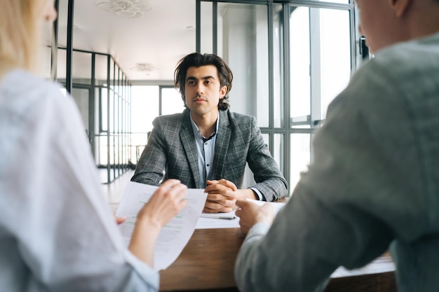 Porträt eines selbstbewussten männlichen Arbeitssuchenden in formeller Kleidung, der von zwei nicht erkennbaren Beratern von Personalmanagern bei einem Treffen in der Unternehmenszentrale interviewt wurde