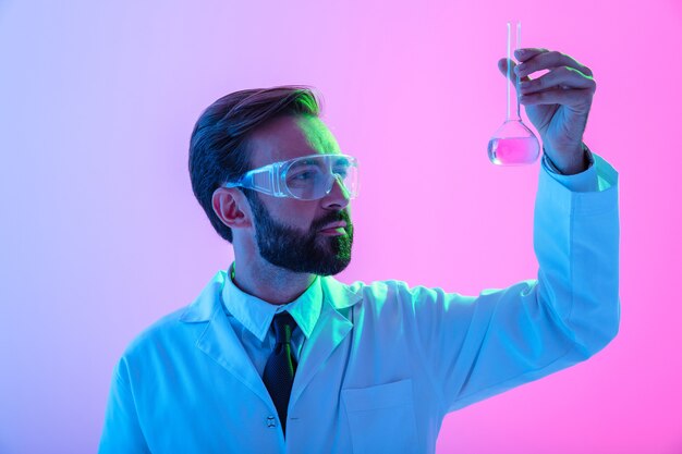 Porträt eines selbstbewussten Wissenschaftlers mit Unifrom und Brille, der isoliert über blau-rosa Dunst steht mit Blick auf eine Flasche mit Flüssigkeit