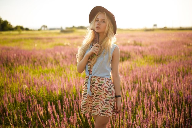 Porträt eines sinnlichen blonden Mädchens mit Hut, das auf einem Blumenfeld auf Sonnenuntergangshintergrund posiert