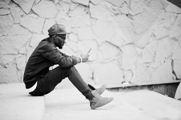 Porträt eines stilvollen afroamerikanischen mannes mit sportswear-mütze und brille, der mit dem telefon zur hand auf der treppe sitzt schwarze männer modellieren straßenmode