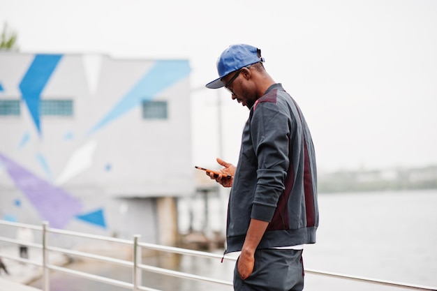 Porträt eines stilvollen afroamerikanischen Mannes mit Sportswear-Mütze und Brille mit Smartphone zur Hand Schwarze Männer modeln auf der Straße gegen den See