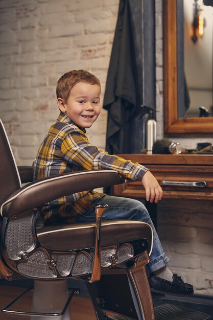 Porträt eines stilvollen kleinen Jungen in Hemd und Jeans, der auf einem Stuhl gegen den Arbeitsplatz des Friseurs sitzt, lächelt und vor der Kamera posiert