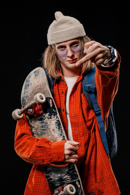 Porträt eines stilvollen Mannes, der im Studio ein Skateboard hält. Nahaufnahme eines lächelnden Skateboarders, der auf schwarzem Hintergrund posiert.