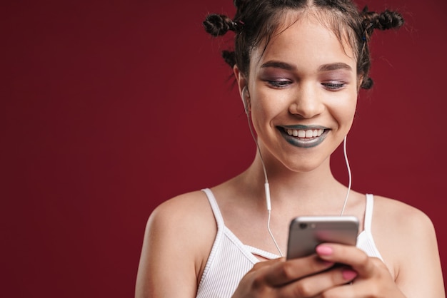 Porträt eines stilvollen Punk-Mädchens mit bizarrer Frisur und dunklem Lippenstift, das lächelt, während es Smartphone mit Kopfhörern verwendet, die über roter Wand isoliert sind?