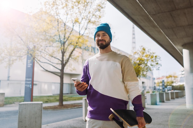 Porträt eines stylischen Skaters hält ein Skateboard in der Hand und geht spazieren. Mann hält Smartphone in der Hand Foto in bester Qualität