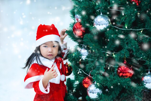 Porträt eines süßen asiatischen kleinen Mädchens, das ein Weihnachtskleid trägt, das glücklich in der Nähe des Weihnachtsbaums istThailand-Kind nimmt am Weihnachtsfest teil