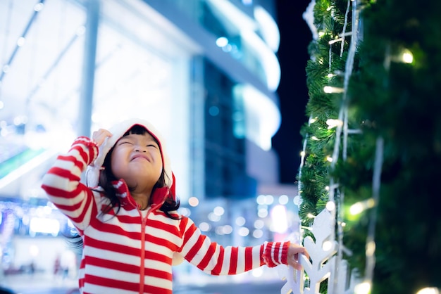 Porträt eines süßen asiatischen kleinen Mädchens, das eine Weihnachtsmütze trägt, die in der Nähe des Weihnachtsbaums glücklich ist