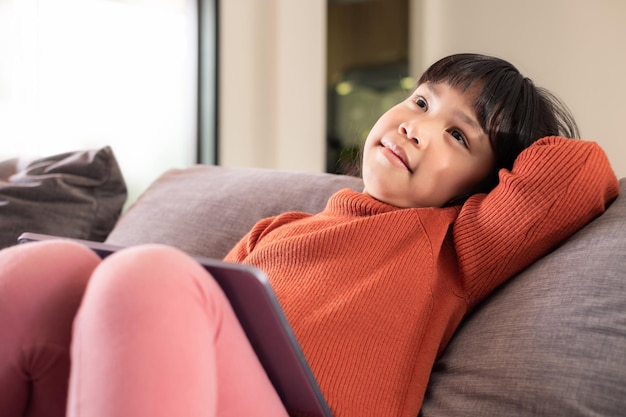 Porträt eines süßen asiatischen Mädchens, das ein Tablet für die Bildung und Einstellung auf einem grauen Sofa in der Nähe des Fensters zu Hause verwendet. Fernunterricht zu Hause Konzept