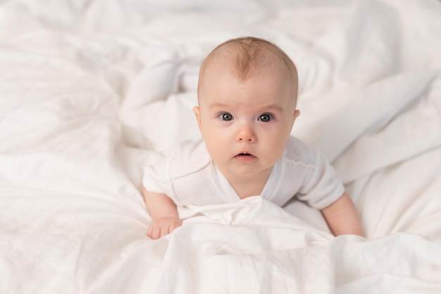 Porträt eines süßen Babys in einem weißen Body auf einem Bett zu Hause mit weißer Bettwäsche. Neugeborenes Baby zu Hause