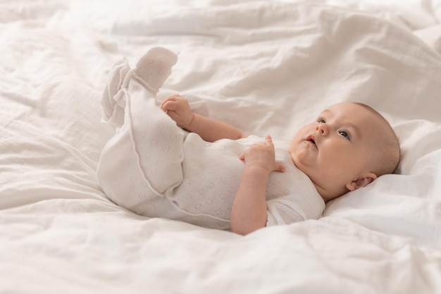 Porträt eines süßen Babys in einem weißen Body auf einem Bett zu Hause mit weißer Bettwäsche. Neugeborenes Baby zu Hause