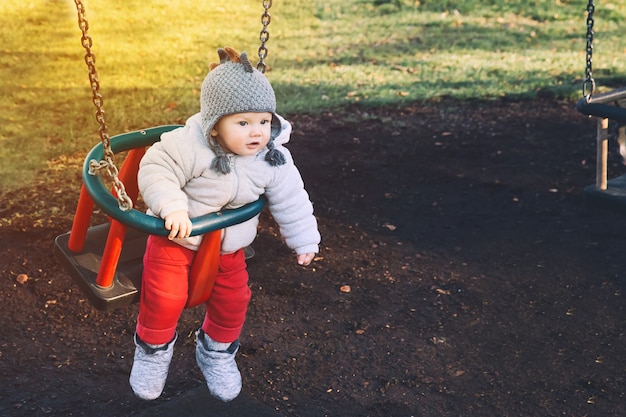 Porträt eines süßen Babys in warmer Kleidung auf der Schaukel auf dem Spielplatz an einem sonnigen Tag im Freien.