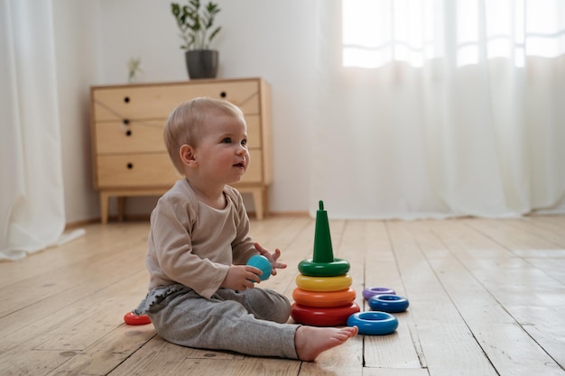 Porträt eines süßen, glücklichen Babykindes, das buntes Spielzeug hält und Pyramidenspiel spielt, das auf dem Boden sitzt