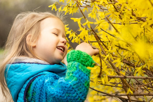 Porträt eines süßen, glücklichen kleinen blonden Mädchens in blauer Jacke in der Nähe von Busch, der mit gelben Blumen blüht