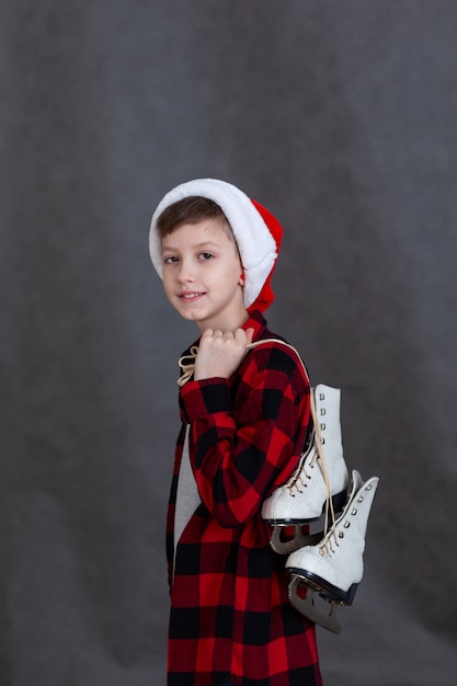 Porträt eines süßen Jungen in einer Weihnachtsmütze und einem roten karierten Hemd, der im Studio auf grauem Hintergrund Schlittschuhe auf seiner Schulter hält.