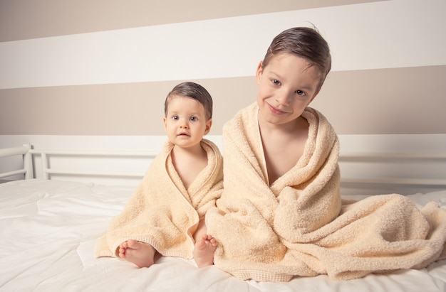 Porträt eines süßen Jungen und eines kleinen Mädchens unter den Handtüchern, die nach dem Bad in einem Bett sitzen