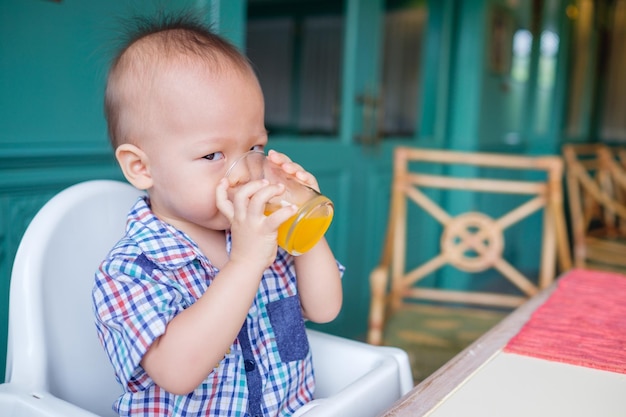 Porträt eines süßen kleinen asiatischen 18 Monate alten Babys, das Fruchtsaft in einem Glas trinkt Lächelnder Junge sitzt im Hochstuhl im Restaurant-Café-Hotel und hält ein Glas leckeren Orangensaft