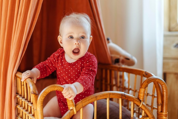Porträt eines süßen kleinen Kindes in roter Babykleidung, das zu Hause an seinem Bett steht