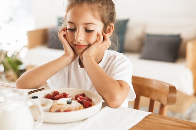 Porträt eines süßen kleinen Mädchens, das am Tisch in der hellen Küche sitzt, während es frühstückt und Pfannkuchen mit Himbeere isst