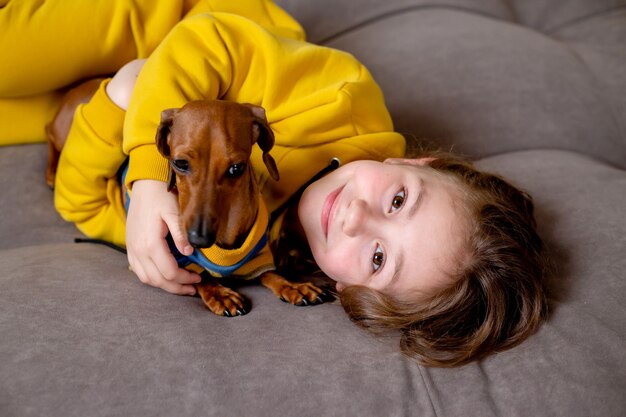 Porträt eines süßen kleinen Mädchens in gelber Kleidung, das mit einem Zwergdackel im blauen Overall im Bett liegt