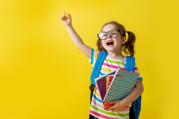 Porträt eines süßen kleinen Mädchens mit Brille in einem gestreiften T-Shirt mit Notizbüchern und Lehrbüchern in den Händen und einem Rucksack. Konzept der Bildung. Fotostudio, gelber Hintergrund, Platz für Text.