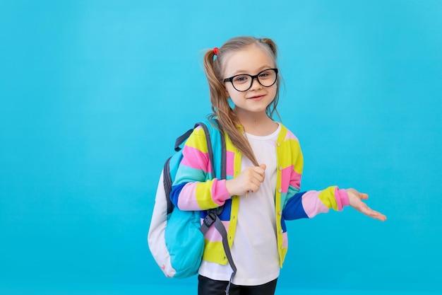 Porträt eines süßen kleinen Mädchens mit Brille in einer gestreiften Jacke und einem Rucksack. Das Konzept der Bildung. Fotostudio, blauer Hintergrund, Platz für Text