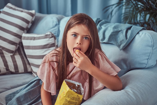 Porträt eines süßen kleinen Mädchens mit langen braunen Haaren, das auf einem Sofa sitzt und Chips isst.