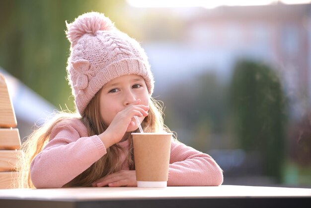 Porträt eines süßen kleinen Mädchens mit rosa Hut, das allein im Straßencafé sitzt und Tee aus Pappbechern trinkt