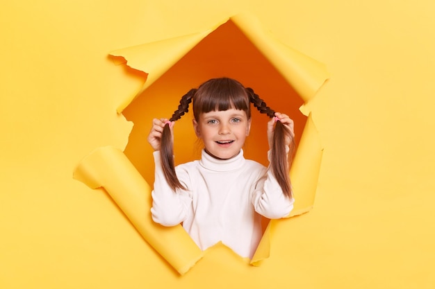 Porträt eines süßen kleinen Mädchens mit Zöpfen, das einen weißen Rollkragenpullover trägt und in einem zerrissenen Loch einer gelben Papierwand posiert, weibliches Kind, das ihre Zöpfe hält und lächelnd in die Kamera schaut