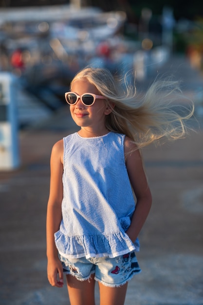 Porträt eines süßen lächelnden kleinen Mädchens mit Brille. Ein Mädchen in kurzen Hosen und einem blauen T-Shirt bei Sonnenuntergang am Meer