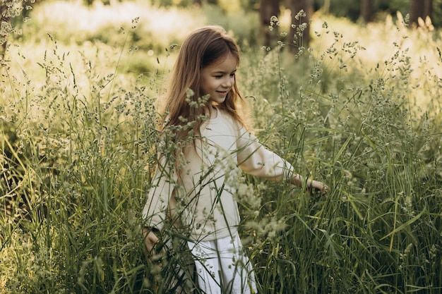 Porträt eines süßen lächelnden Mädchens mit schönen Haaren, das für ein Foto inmitten von hohem Gras in einem Nadelwald bei Sonnenuntergang posiert
