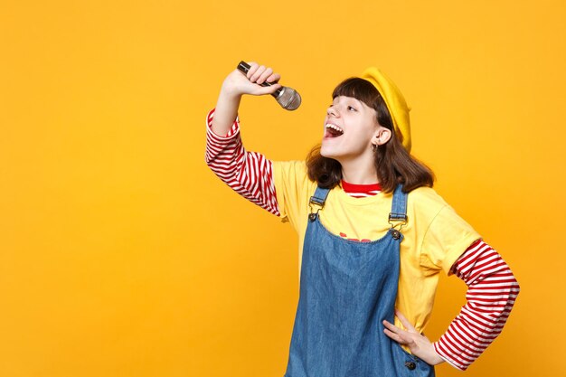 Porträt eines süßen Mädchen-Teenagers in französischer Baskenmütze und Denim-Sommerkleid singt Lied im Mikrofon isoliert auf gelbem Wandhintergrund im Studio. Menschen aufrichtige Emotionen, Lifestyle-Konzept. Mock-up-Kopienbereich.