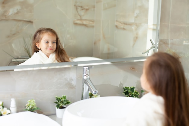 Porträt eines süßen Mädchens, das in einem weißen Mantel mit einem Einhorn in einem hellen Badezimmer mit Fenster in den Spiegel schaut. Hygiene. Morgenroutine. Reinheit. Wie Mutter. Foto in hoher Qualität