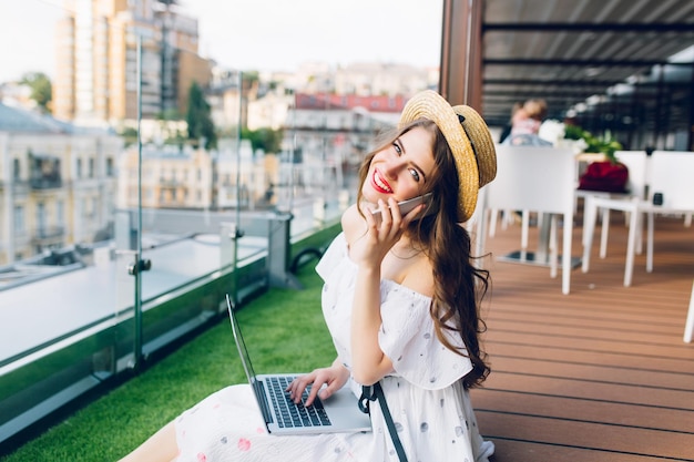 Porträt eines süßen Mädchens mit langen Haaren im Hut, das auf dem Boden auf der Terrasse sitzt. Sie trägt ein weißes Kleid mit nackten Schultern, roten Loipenstab. Sie tippt auf Laptop und telefoniert.