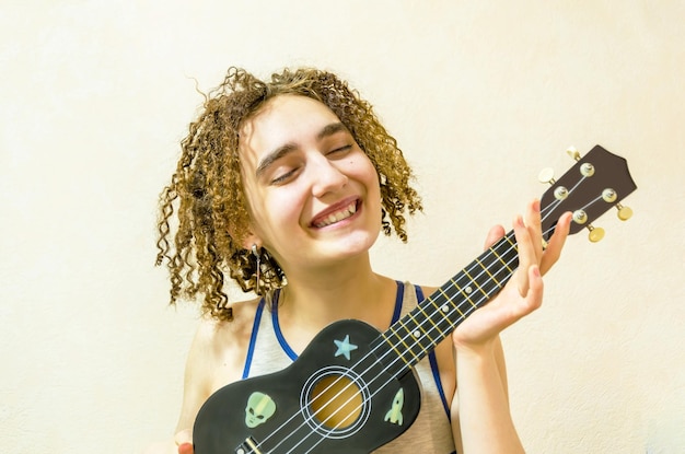 Porträt eines süßen Mädchens mit zerzausten lockigen Haaren, das Ukulele spielt. Junge lächelnde Frau mit Ukulele