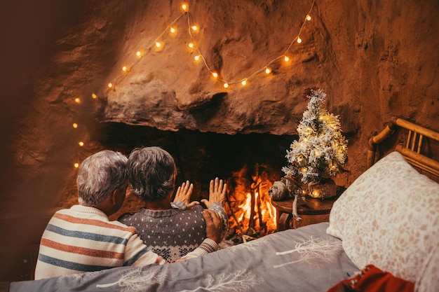 Porträt eines süßen, reifen Seniors, der Weihnachten vor dem heißen Kamin zu Hause genießt und sich kalt umarmt und die Hände hält, die farbige Urlaubssocken in feetxA tragen