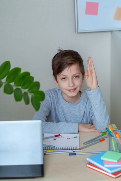 Porträt eines süßen Schuljungen, der zu Hause an einem Tisch vor einem Computer sitzt und seine Hand hebt. Zurück zur Schule. Online-Bildung. Kind macht Hausaufgaben