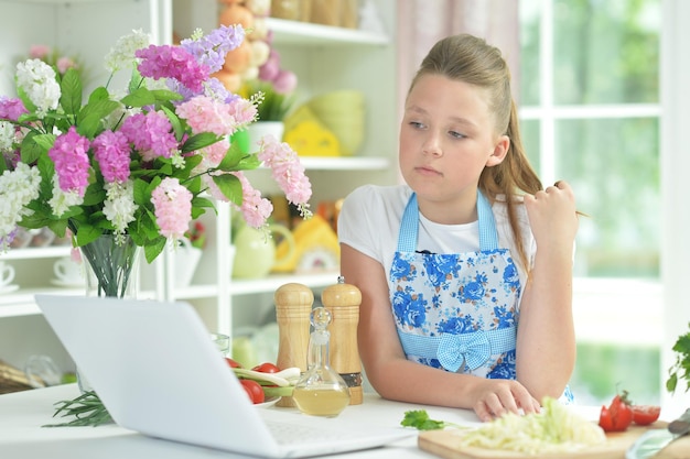Porträt eines süßen Teenager-Mädchens mit Laptop, während frischer Salat auf dem Küchentisch zubereitet wird