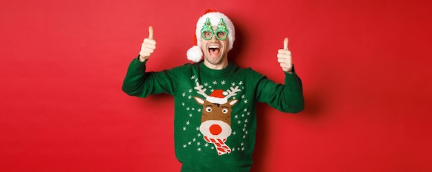 Foto porträt eines superglücklichen jungen mannes in partybrille, weihnachtsmütze und pullover, daumen hoch zur zustimmung zeigend, neujahrs-promo-angebot empfehlend, auf rotem hintergrund stehend.