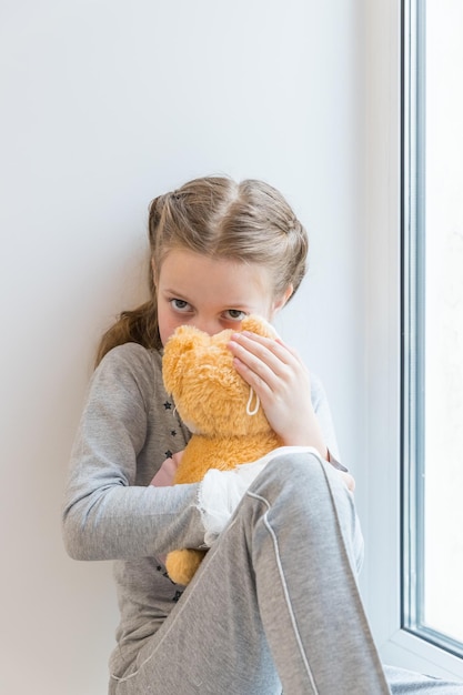 Porträt eines traurigen Kindes, das mit einem Stofftier auf der Fensterbank sitzt junges Mädchen ist mit Teddybär im Krankenhaus...