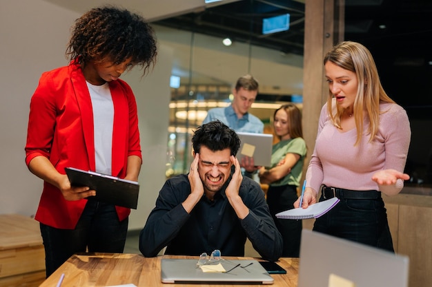 Porträt eines überarbeiteten Teamleiters unter Stress, der mit einem Laptop mit Schmerzkopf am Tisch sitzt Multiethnische Kollegen schütteln Dokumente, die in der Nähe stehen