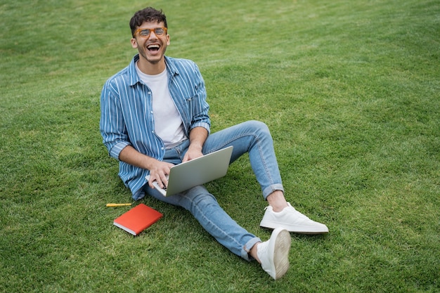 Porträt eines überglücklichen indischen freiberuflers, der einen laptop verwendet, der online auf gras sitzt