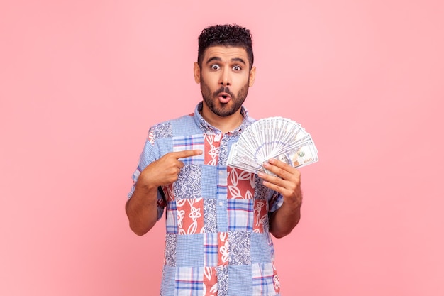 Porträt eines überraschten bärtigen Mannes, der auf einen Fan von Dollarbargeld zeigt, Geldwechsel, schockiert über den Zinssatz, der mit Erstaunen in die Kamera blickt. Studioaufnahme im Innenbereich isoliert auf rosa Hintergrund.