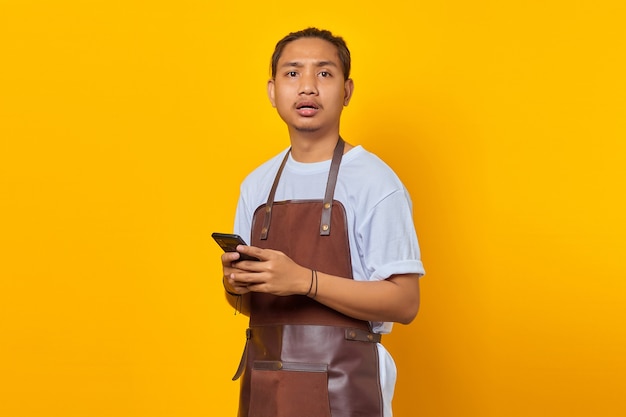 Porträt eines überraschten, gutaussehenden Barista, der nach vorne schaut und Smartphone auf gelbem Hintergrund hält