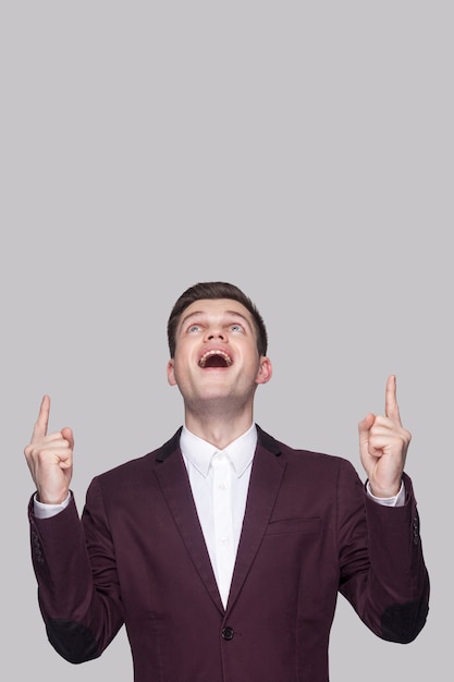 Porträt eines überraschten, gutaussehenden jungen Mannes in violettem Anzug und weißem Hemd, stehend, nach oben schauend und mit erstauntem Gesicht auf den Kopierraum oben zeigend. Indoor-Studioaufnahme, auf grauem Hintergrund isoliert