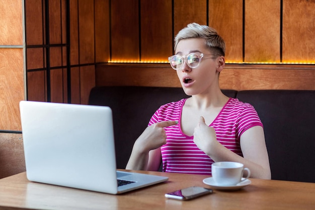 Porträt eines überraschten jungen Mädchens mit blonden Haaren in rosafarbenem T-Shirt sitzt allein im Café und hat einen neuen Vorschlag für eine Arbeit am Laptop, der mit großen Augen und offenem Mund mit dem Finger auf sich selbst zeigt