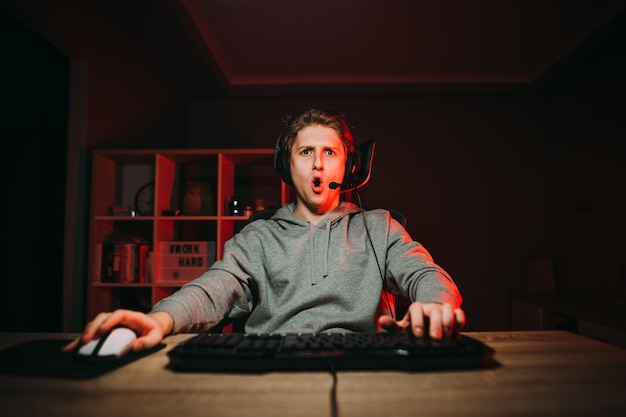 Porträt eines überraschten jungen männlichen Spielers in Freizeitkleidung und Headset, der in einem Raum sitzt