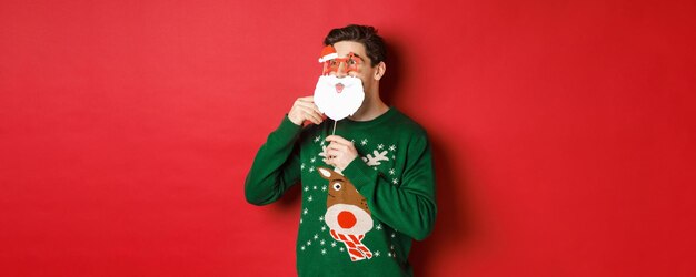 Porträt eines überraschten Mannes im grünen Weihnachtspullover, der eine lustige Weihnachtsmann-Maske hält, das neue Jahr feiert, verblüfft nach links schaut und über rotem Hintergrund schleift