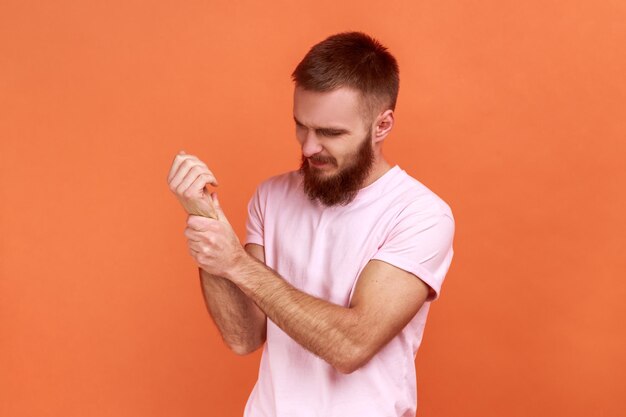 Porträt eines ungesunden bärtigen mannes, der mit offenem mund steht, schmerz fühlt, wundes handgelenk massiert, handverletzungen erleidet, rosa t-shirt trägt. Studioaufnahme im Innenbereich isoliert auf orangefarbenem Hintergrund.