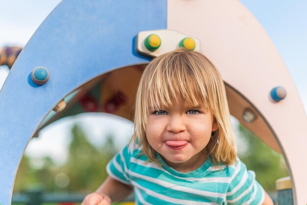 Porträt eines ungezogenen kleinen Jungen, der die Kamera betrachtet, die seine Zunge auf dem Spielplatz herausstreckt.